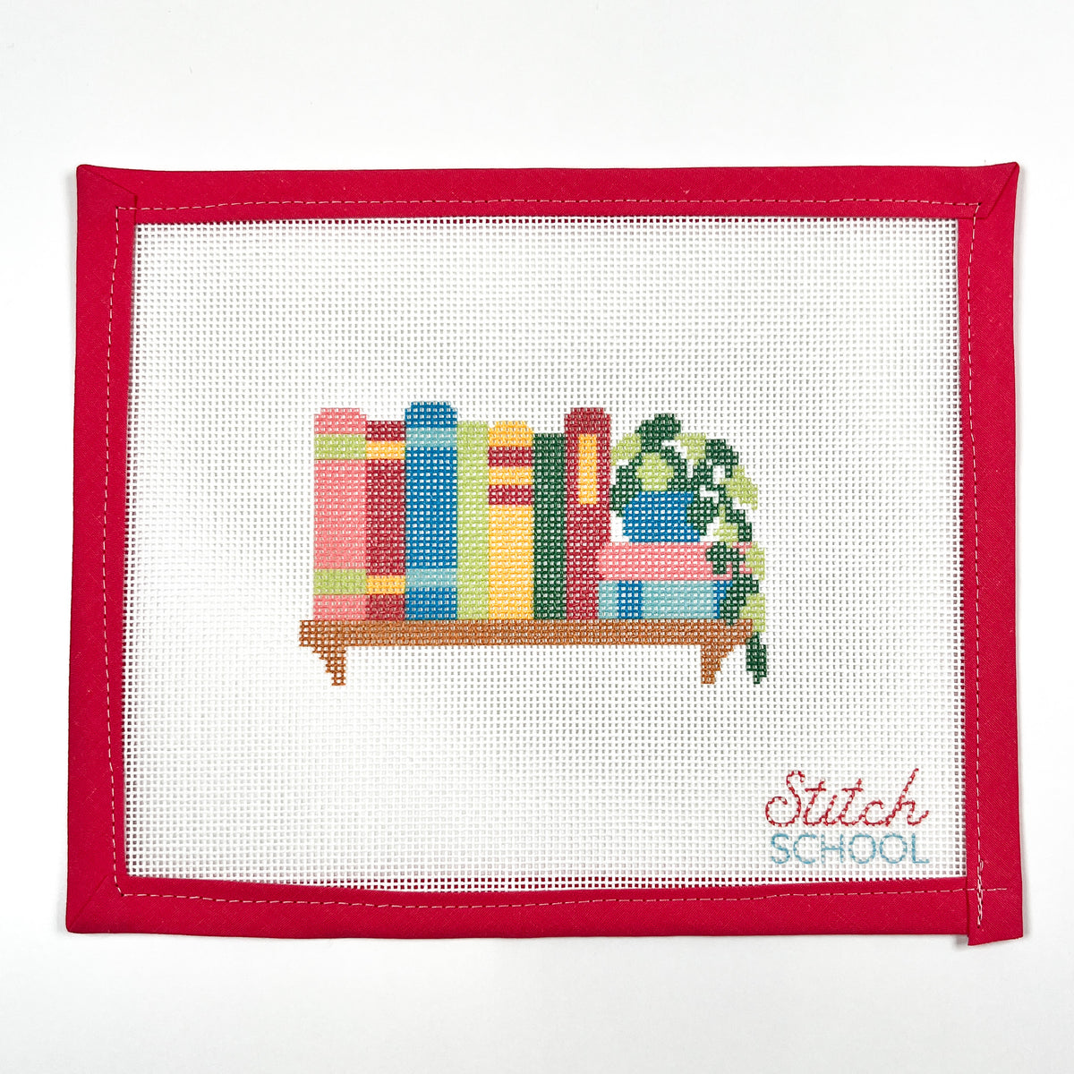 Happy Little Bookshelf needlepoint canvas with colorful books and a trailing plant on a brown shelf, shown with red binding.
